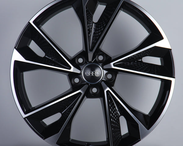 A polished, multi-spoke car wheel with a metallic finish, showcasing the Audi emblem in its center, is displayed against a plain gray background.