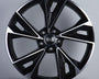 A black and silver alloy wheel is displayed upright, featuring a modern design with multiple spokes. The "Audi" logo is prominently centered. The background is a simple, neutral gray.