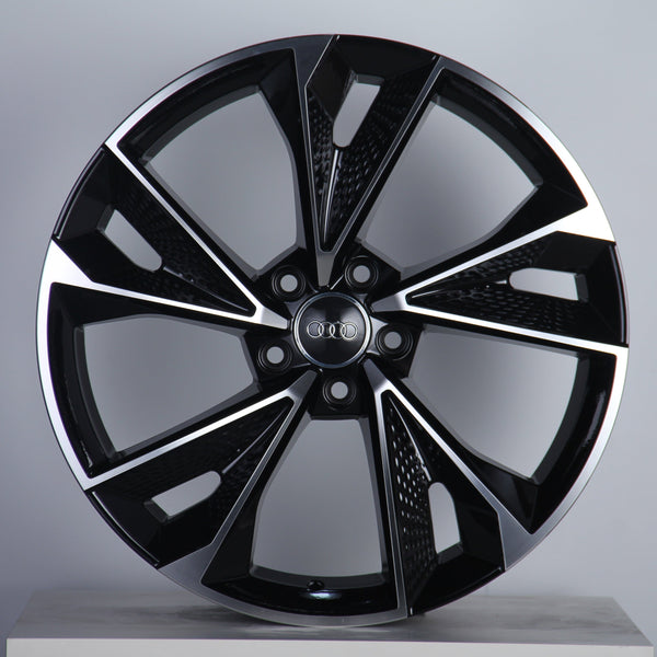 A black and silver alloy wheel is displayed upright, featuring a modern design with multiple spokes. The "Audi" logo is prominently centered. The background is a simple, neutral gray.