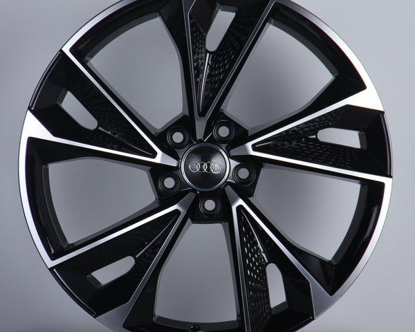 A black and silver alloy wheel with a modern, angular design is standing upright. The center features the Audi logo with interlocking rings. The background is a plain, light gray surface.