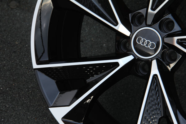 A black and silver Audi car wheel with an intricate design is stationary on a textured asphalt surface. Four interlinked rings are centered on the hubcap.