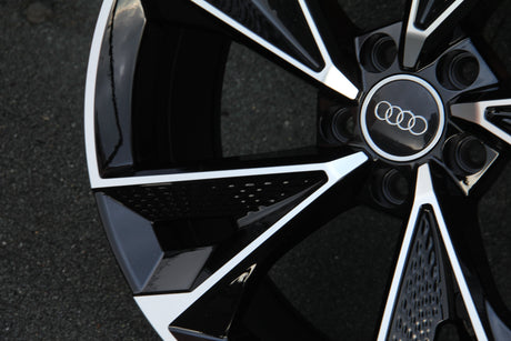 A shiny black and silver Audi wheel, stationary, is shown close-up on a textured, gray asphalt surface. Four interlocking rings are centered.
