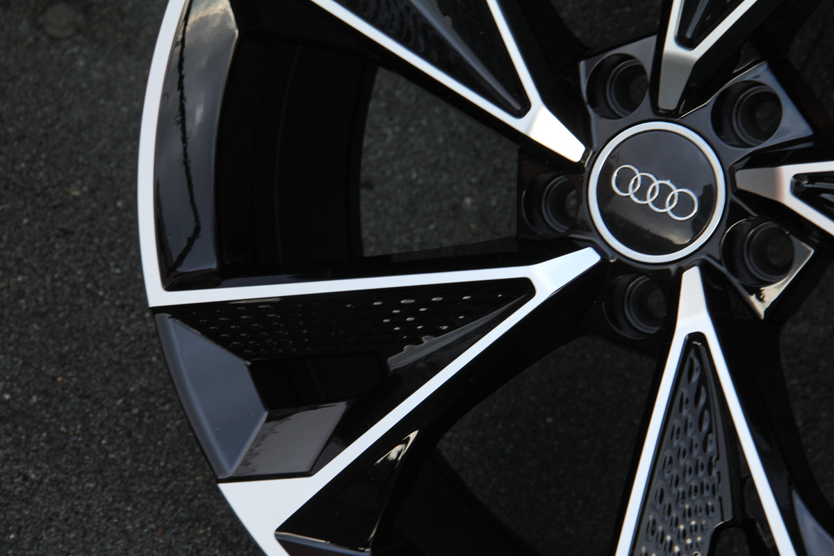 A stylish black and silver car wheel rim with the Audi logo in the center, resting on a textured asphalt surface.