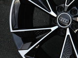 A glossy black Audi wheel rim with a silver logo sits stationary against a textured asphalt background, featuring a complex angular design with contrasting white accents.