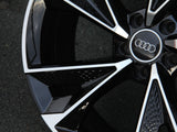 A shiny black and silver car wheel rim with the Audi logo at the center, positioned on a dark, textured ground surface.