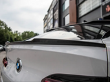 A white BMW with a prominent rear spoiler, parked on a street with trees and modern buildings in the background.