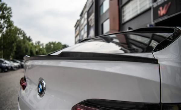 A white BMW with a prominent rear spoiler, parked on a street with trees and modern buildings in the background.