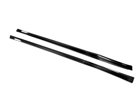 Two sleek, black carbon fiber side skirts lie parallel on a white background, showcasing a streamlined design with smooth, reflective surfaces and tapering ends.