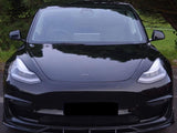 A black car, front view, parked on a road with a background of green trees and grass. The car has sleek headlights, a shiny exterior, and a logo on the hood.