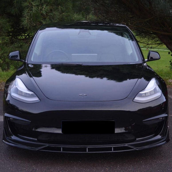 A black car, front view, parked on a road with a background of green trees and grass. The car has sleek headlights, a shiny exterior, and a logo on the hood.