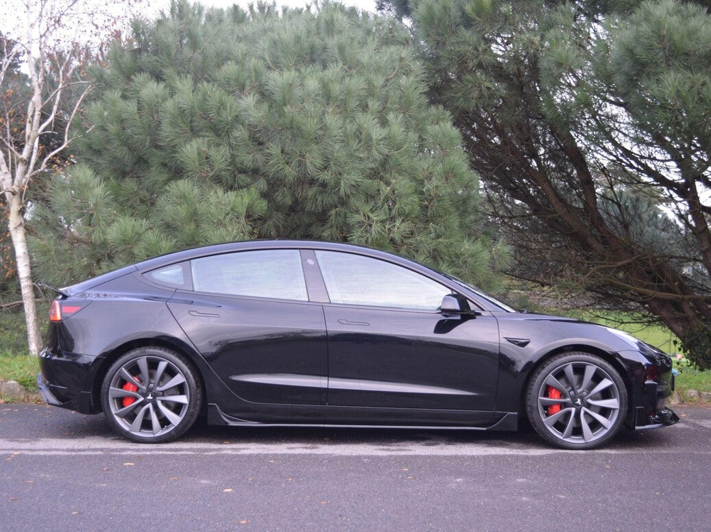 A black car is parked on a paved surface, featuring red brake calipers and large wheels. It is situated next to lush, green pine trees and a grassy area.