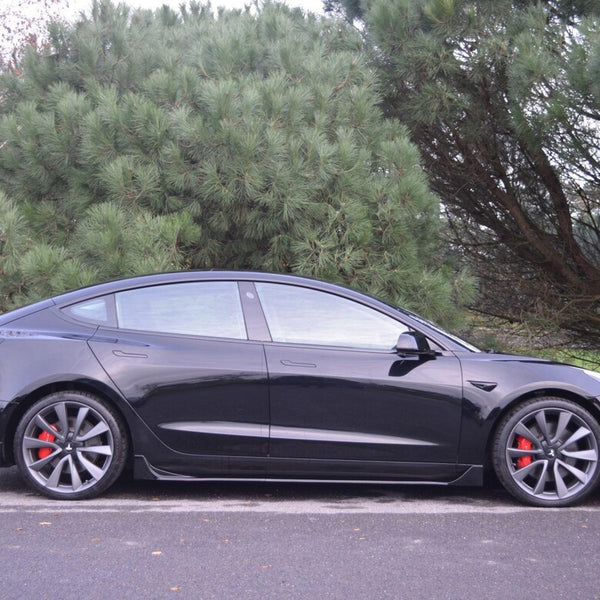 A black car is parked on a paved surface, featuring red brake calipers and large wheels. It is situated next to lush, green pine trees and a grassy area.