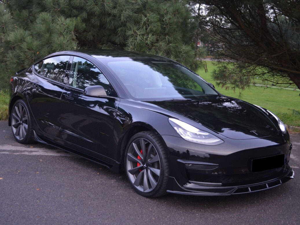 A black sedan with large wheels and bright headlights is parked on a paved road. It's surrounded by lush green trees, suggesting a park or wooded area.