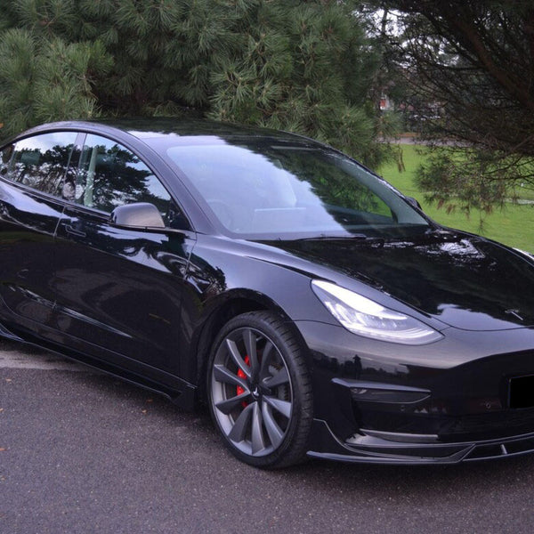 A black sedan with large wheels and bright headlights is parked on a paved road. It's surrounded by lush green trees, suggesting a park or wooded area.