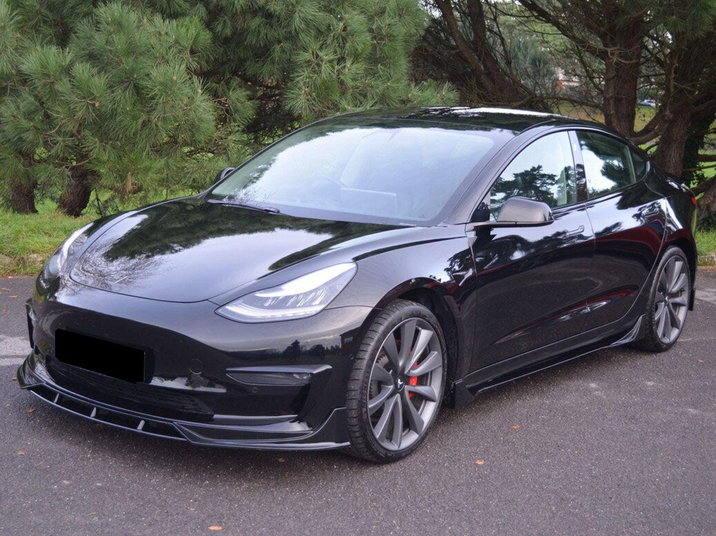 A sleek black car is parked on a paved area, surrounded by lush green trees, showcasing its streamlined, modern design and alloy wheels.