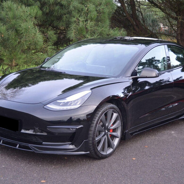 A sleek black car is parked on a paved area, surrounded by lush green trees, showcasing its streamlined, modern design and alloy wheels.