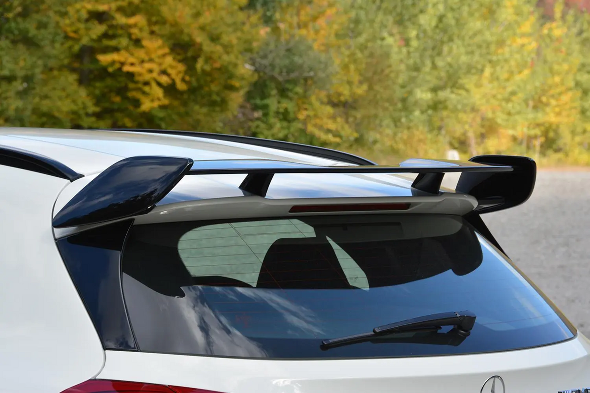 A sleek car spoiler perched on the rear of a white vehicle, situated against a backdrop of green and yellow foliage on a clear day.