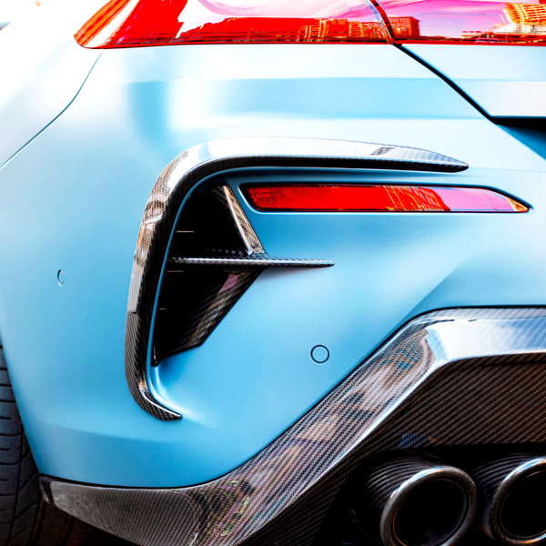 A blue sports car with a carbon fiber rear bumper features sleek, dual exhaust pipes and vibrant red taillights, parked outdoors.