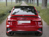 A red Audi S9 is parked on a narrow dirt road flanked by tall trees and lush greenery, under a cloudy sky.