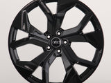 A glossy black car wheel rim with a modern design features five split spokes. In the center, four interlocking rings in silver logo represent Audi, against a plain white background.