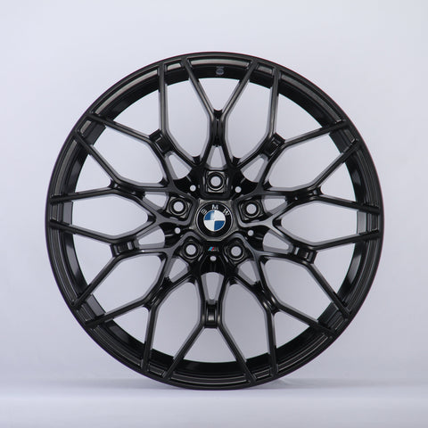 A black alloy wheel with a complex, multi-spoke design is displayed against a plain white background. The center features the BMW logo, surrounded by intricate patterns.