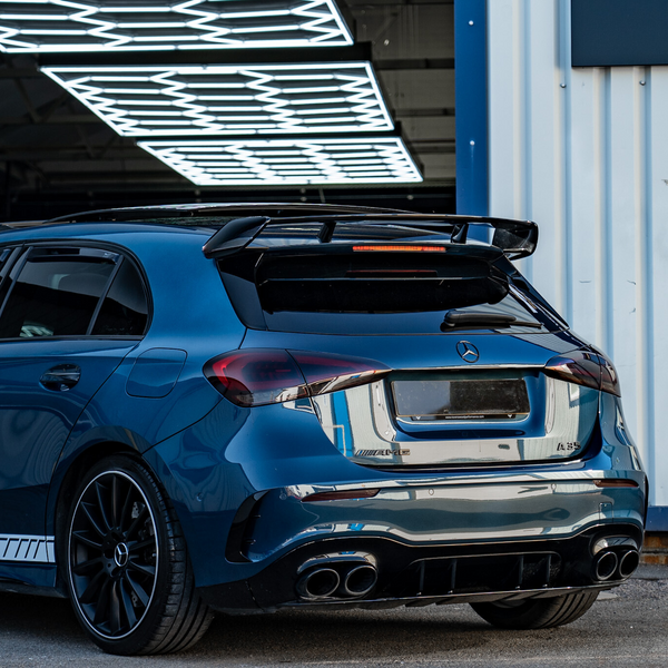 A blue sports car is parked outside a garage with neon lights above. The car has "AMG" branding and features a rear spoiler and quad exhaust pipes.