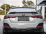A silver BMW 4 Series (M440i) car is parked on a street, featuring dual exhaust pipes and a blurred license plate. Surrounded by trees and buildings under a cloudy sky.
