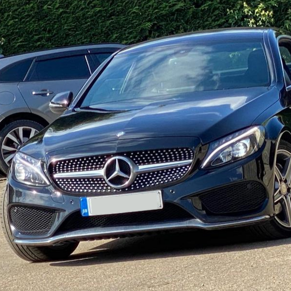 A black Mercedes-Benz car, parked on a street, with a grill displaying the emblem, beside another vehicle and green shrubbery in the background.