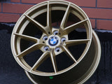 A gold BMW car wheel rim rests upright on a dark surface against a brick wall, displaying the BMW logo at its center.