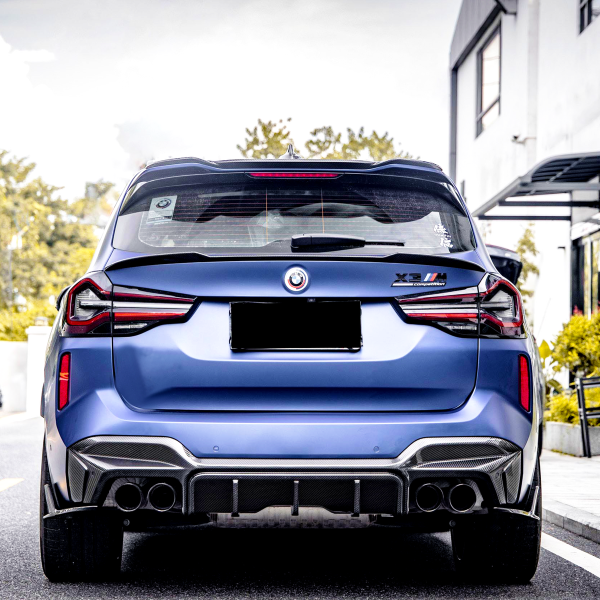 A blue BMW X3 M with a license plate concealed, parked outdoors. The badges on the back indicate “X3 M Competition.” Nearby are buildings and greenery.