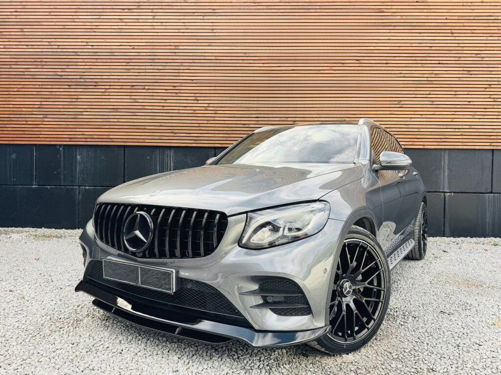 A sleek, metallic gray SUV with a prominent grille and large black wheels is parked on gravel, set against a wooden and black paneled wall.