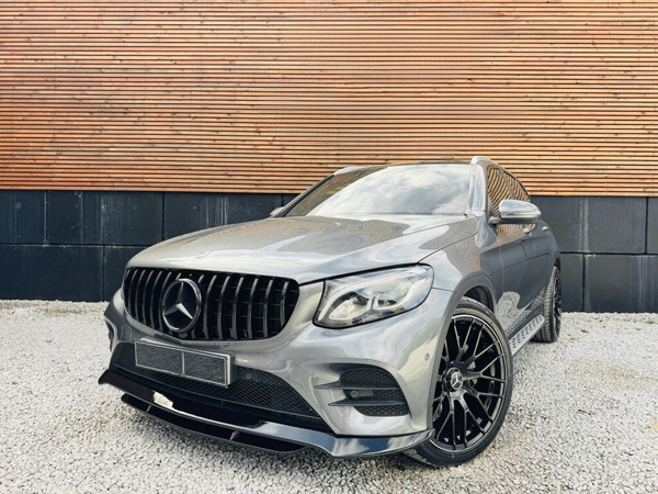 A sleek, metallic gray SUV with a prominent grille and large black wheels is parked on gravel, set against a wooden and black paneled wall.