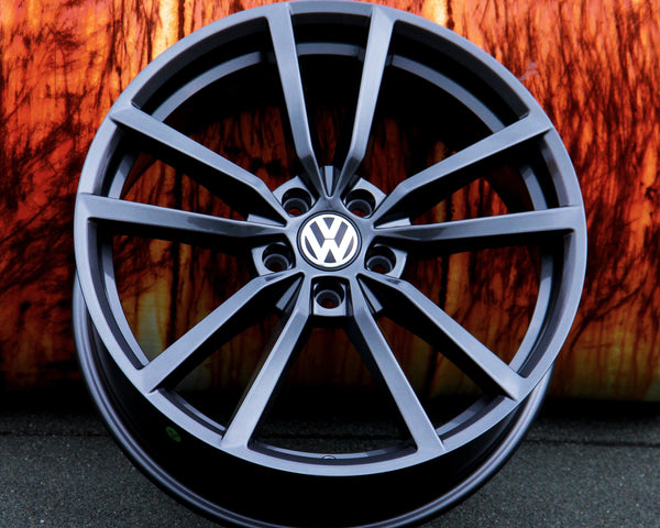 A black alloy wheel with the Volkswagen logo is propped against a textured, rust-colored background, resting on a dark asphalt surface.