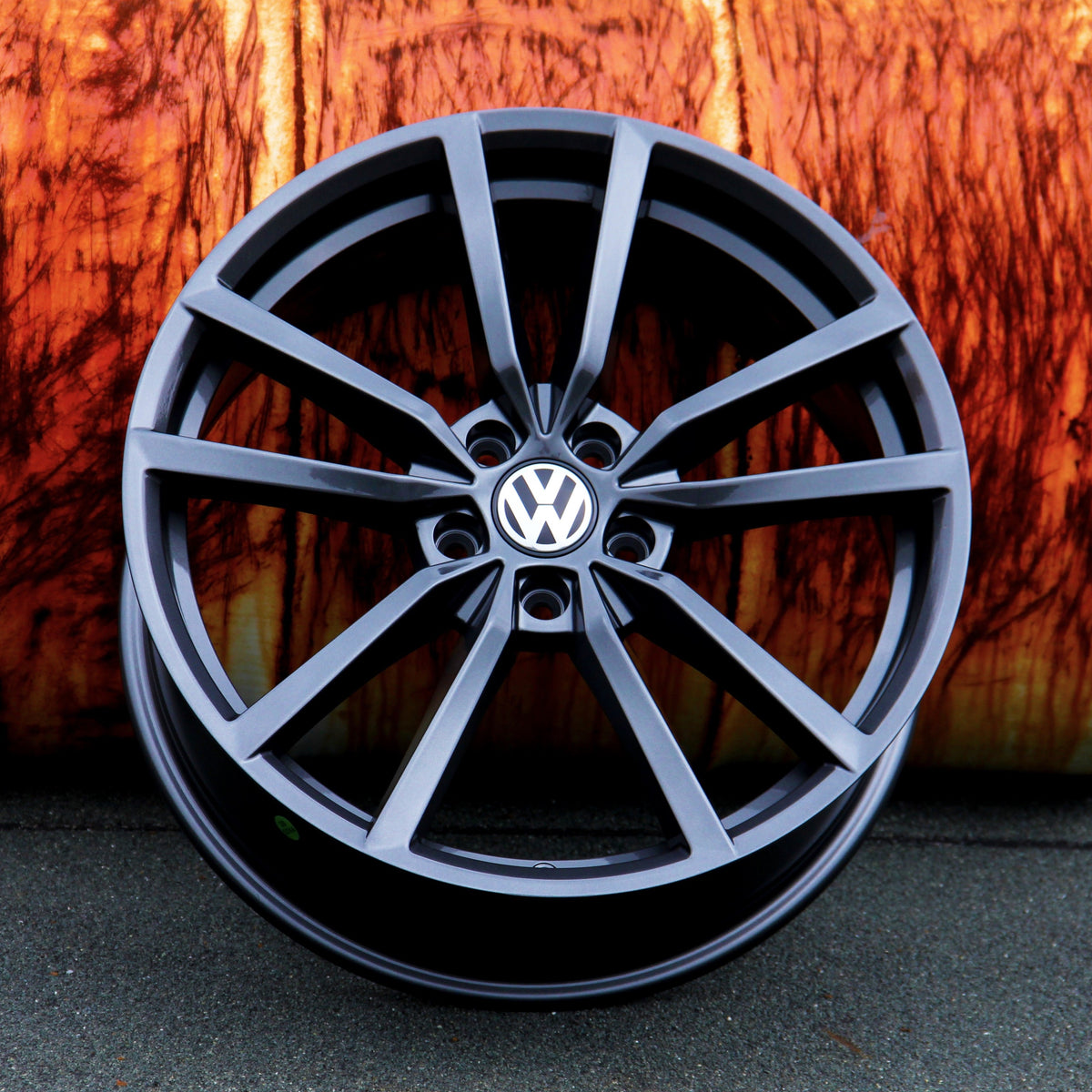 A black alloy wheel with the Volkswagen logo is propped against a textured, rust-colored background, resting on a dark asphalt surface.