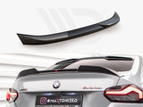 A glossy black rear spoiler sits unattached above a grey BMW M240i in a showroom. Text on the license plate reads 