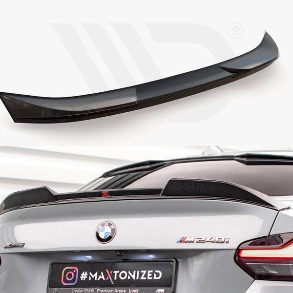 A glossy black rear spoiler sits unattached above a grey BMW M240i in a showroom. Text on the license plate reads "#MAXTONIZED" with an Instagram logo.