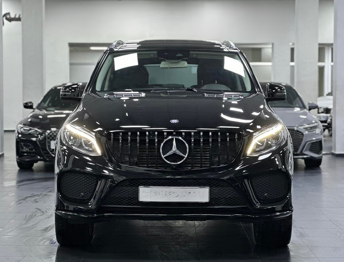 A black Mercedes-Benz SUV stands prominently with headlights on, inside a showroom, flanked by two other black cars in the background.
