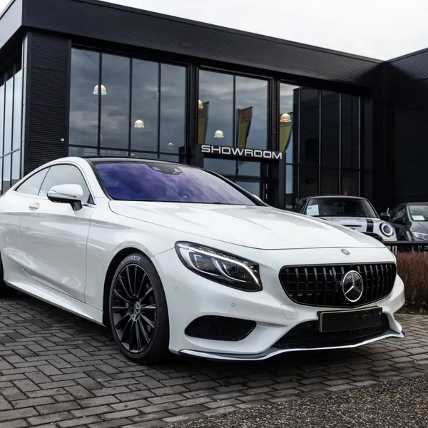 A white luxury car is parked outside a modern glass-walled showroom, positioned on a cobblestone driveway. The word "SHOWROOM" is visible on the building behind.
