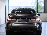 A sleek, dark-colored BMW car parked indoors with quad exhaust pipes, illuminated taillights, and a rear spoiler. Large windows reveal a blurred view outside.