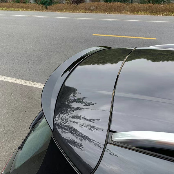 A car's glossy rear spoiler, reflecting nearby trees, is parked beside an asphalt road with white and yellow lane markings. Bushes line the road in the background.