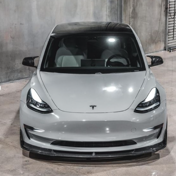 A sleek white car with a glossy finish is parked in an industrial garage with concrete walls. The front features a subtle emblem on the hood.