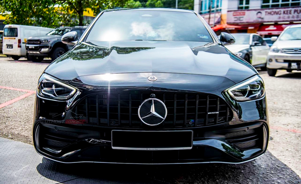 A black Mercedes-Benz AMG car is parked outdoors on a sunny day, surrounded by other vehicles. The brand logo is prominently displayed on the grille.