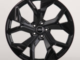 A glossy black alloy wheel with a five-spoke design featuring the Audi logo at the center, sits against a plain white background.
