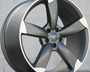 A metallic car wheel rim with five spokes, partially reflective, is propped upright against a plain, light-colored wall. The design features a combination of matte and glossy finishes.