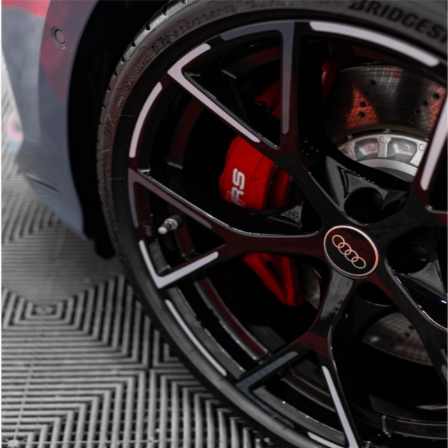 A black car wheel with an Audi logo features a red brake caliper marked "RS." It rests on a textured mat, showcasing a shiny, modern design.