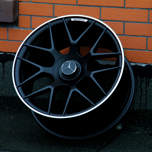 A black alloy wheel with a Mercedes-Benz emblem rests against a brick wall. The text "AMG" is visible on the rim's exterior.