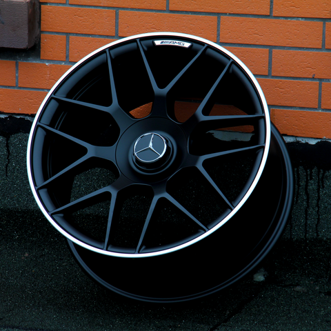 A black alloy wheel with a Mercedes-Benz emblem rests against a brick wall. The text "AMG" is visible on the rim's exterior.