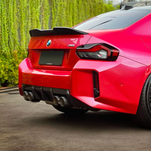 A red BMW M2 car is parked on a driveway, displaying a quad exhaust and black spoiler, with green ivy-covered walls in the background.