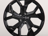 A glossy black alloy wheel with a multi-spoke design. The Audi logo is centered on the hub. It's set against a plain, light grey background.