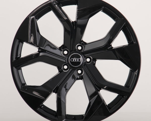 A glossy black alloy wheel with a multi-spoke design. The Audi logo is centered on the hub. It's set against a plain, light grey background.